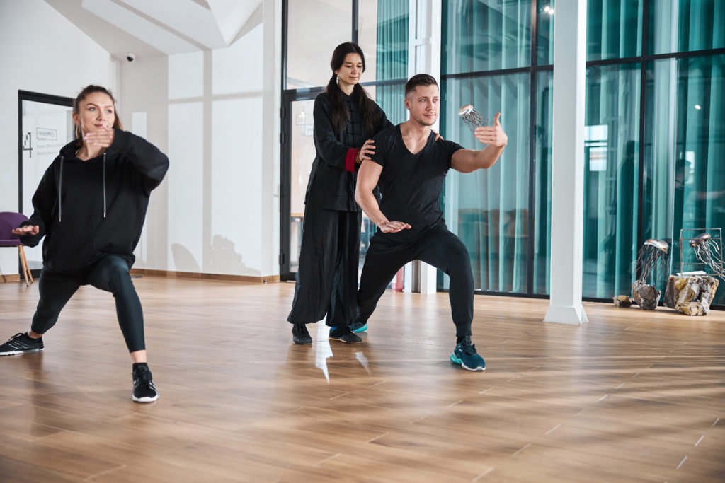 Tai chi master teaching pair of Caucasian students stance while correcting their posture in classroom with steel jellyfish figures