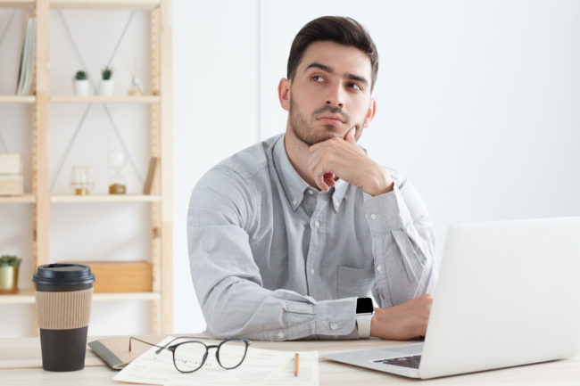Young business man in gray shirt working on laptop in office, looking aside, thinking of new decisions for his company or project