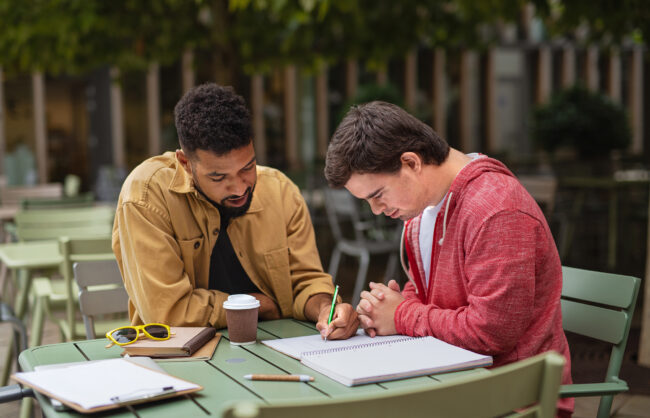 A young man with Down syndrome with his mentoring friend sitting outdoors in cafe and studying.